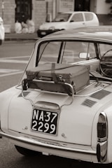 A vintage car with a suitcase strapped to its rear, parked on the street. The scene includes a classic style taillight and a retro license plate. In the background, a blurred view of a parked modern car and a building facade can be seen. The photograph is captured in black and white.