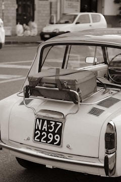 A vintage car with a suitcase strapped to its rear, parked on the street. The scene includes a classic style taillight and a retro license plate. In the background, a blurred view of a parked modern car and a building facade can be seen. The photograph is captured in black and white.