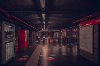 A dimly lit underground metro station hall with ticket machines on the left and a large map on the right. The space is marked with red lines and handles for directing passengers. Ceiling lights run in a straight line, reflecting off the shiny black floor. A few turnstiles are visible, leading to the platform area in the distance. Minimal foot traffic contributes to the quiet and isolated feel of the scene.
