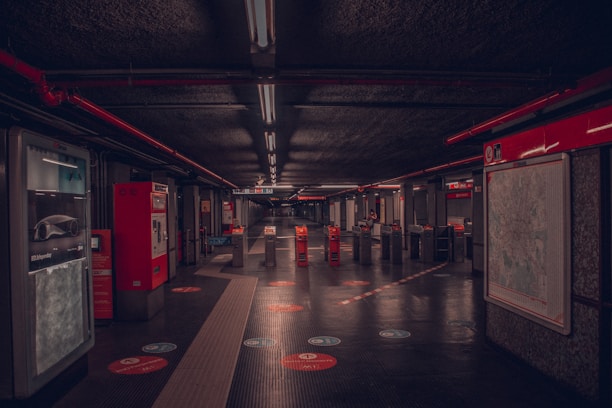 A dimly lit underground metro station hall with ticket machines on the left and a large map on the right. The space is marked with red lines and handles for directing passengers. Ceiling lights run in a straight line, reflecting off the shiny black floor. A few turnstiles are visible, leading to the platform area in the distance. Minimal foot traffic contributes to the quiet and isolated feel of the scene.
