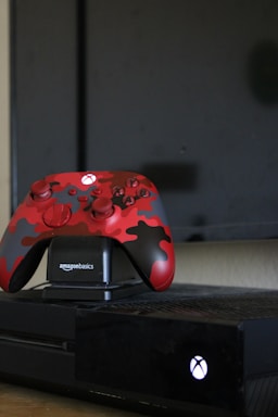 A black gaming console with a red and black camo-patterned controller on an AmazonBasics charging dock. The console and controller are positioned in front of a television screen.