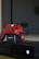 A black gaming console with a red and black camo-patterned controller on an AmazonBasics charging dock. The console and controller are positioned in front of a television screen.