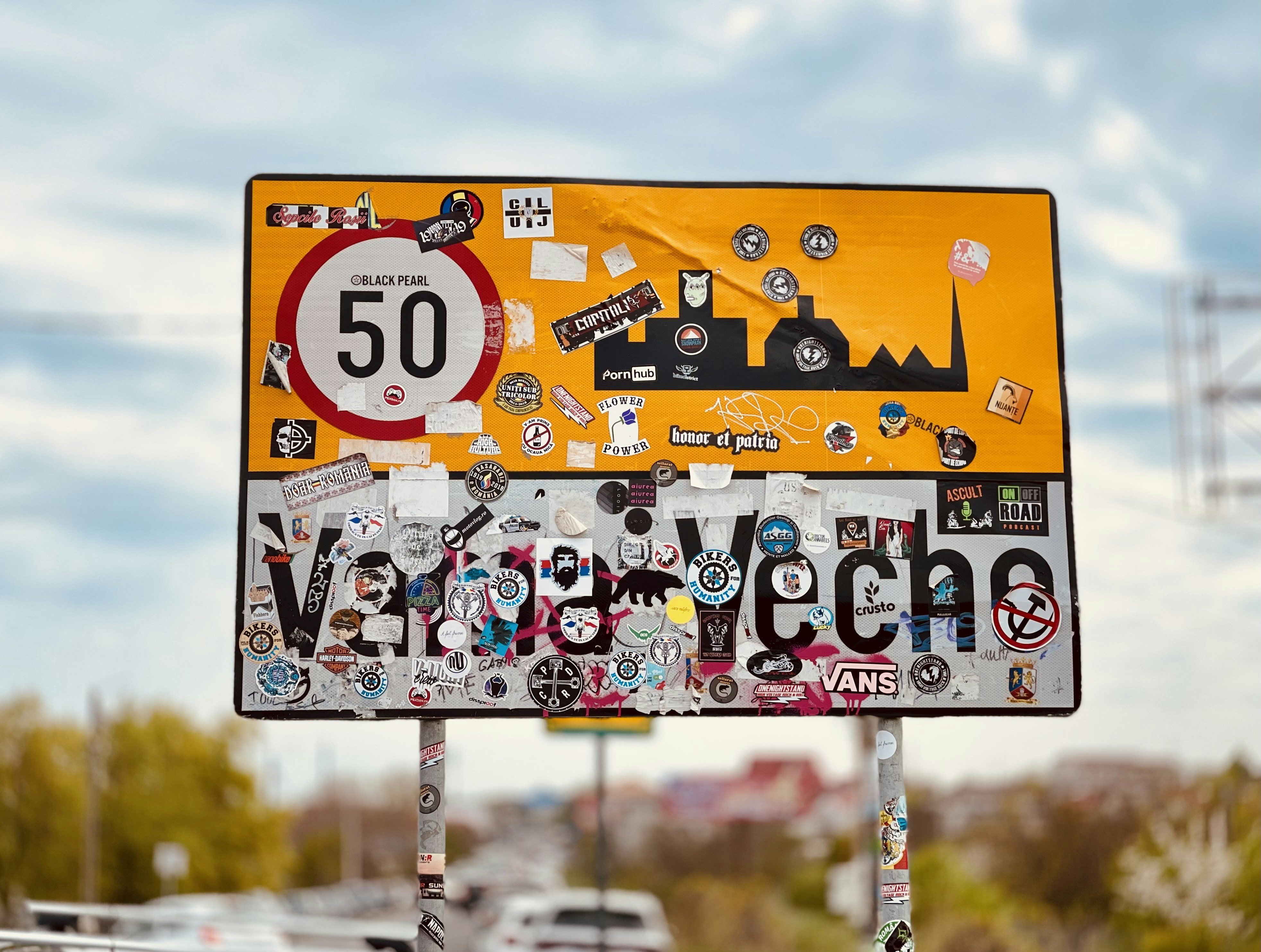 Weathered yellow/white "Vama Veche" entry sign with a 50 limit and skyline. The surface is heavily plastered with colorful stickers, logos, and slogans against a blurred road background.