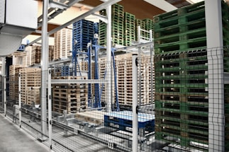 A warehouse area is filled with stacks of wooden and plastic pallets organized on metal racks. The foreground shows a black and white metal fence separating the pallets from the walkway.