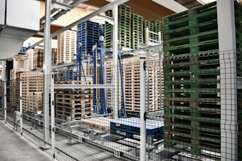A warehouse area is filled with stacks of wooden and plastic pallets organized on metal racks. The foreground shows a black and white metal fence separating the pallets from the walkway.