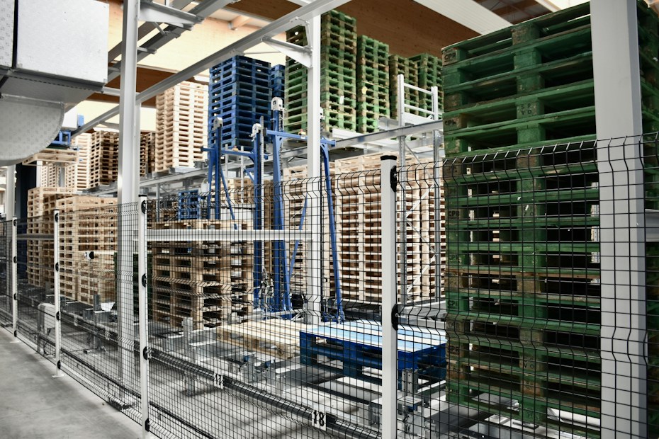 A warehouse area is filled with stacks of wooden and plastic pallets organized on metal racks. The foreground shows a black and white metal fence separating the pallets from the walkway.