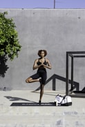 A person is practicing yoga in an outdoor setting, balancing on one leg in a tree pose. They are wearing athletic attire and are positioned on a yoga mat. A large gray wall serves as the backdrop, and a small tree provides some greenery to the left. A duffle bag and a bottle are placed on the ground near the mat.
