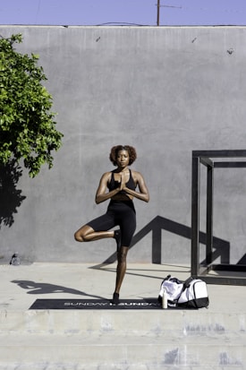 A person is practicing yoga in an outdoor setting, balancing on one leg in a tree pose. They are wearing athletic attire and are positioned on a yoga mat. A large gray wall serves as the backdrop, and a small tree provides some greenery to the left. A duffle bag and a bottle are placed on the ground near the mat.