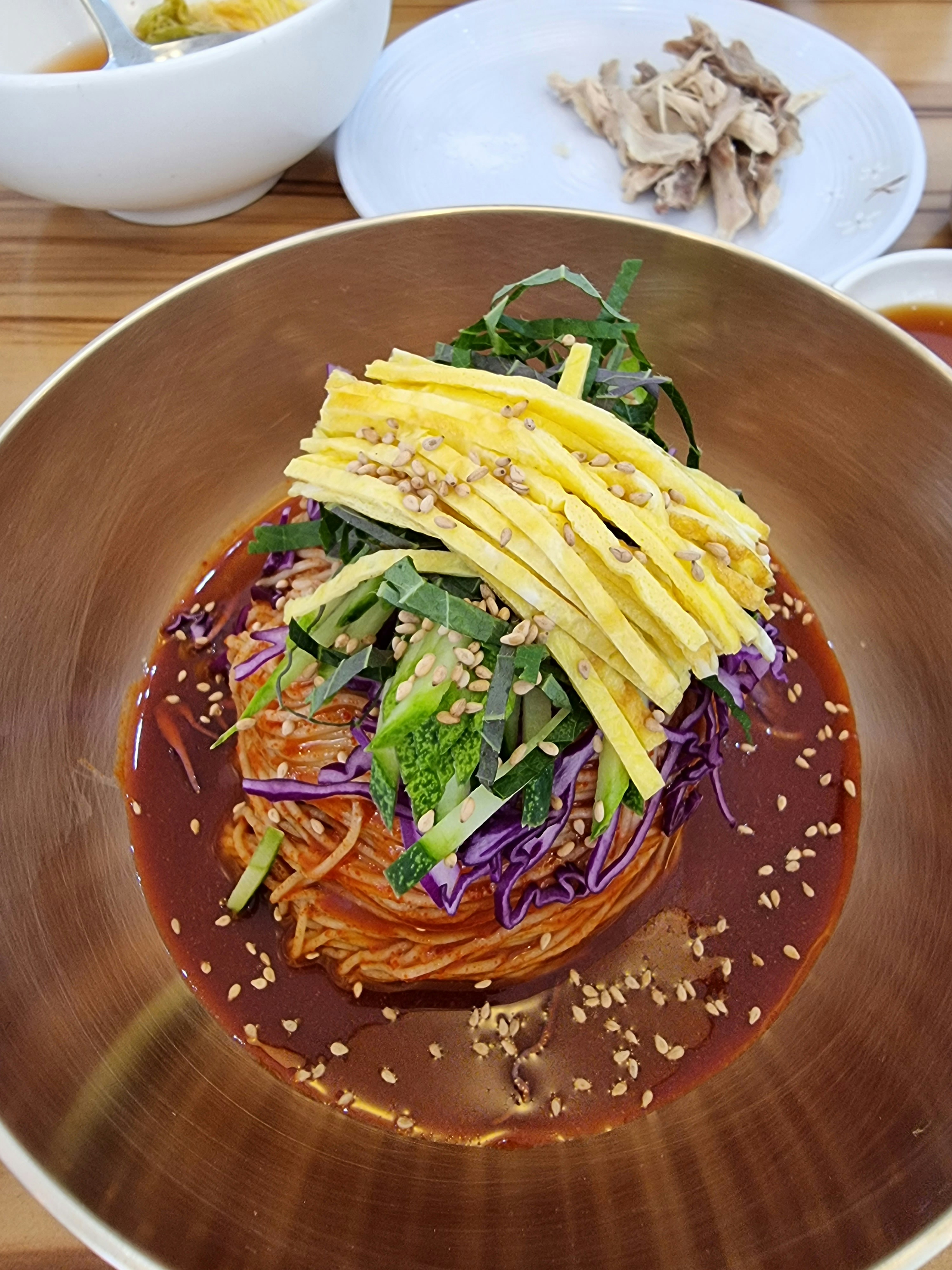 Cold Korean buckwheat noodles naengmyeon in silver bowl with ice, cucumber slices, and half boiled egg