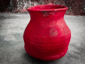 A red clay pot with a wide body and a narrow opening is positioned on a grey concrete surface. It has a rough texture and appears to be hand-painted, with visible brush strokes and uneven patches of color. The background consists of a faded, slightly rough wall with a blend of dark and light patches.