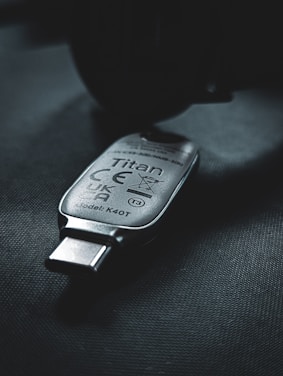 A USB security key marked with the name 'Titan', featuring various certification symbols and model number K40T, lies on a dark surface. The lighting creates a moody atmosphere with emphasis on the metallic texture of the key.