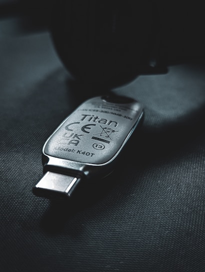 A USB security key marked with the name 'Titan', featuring various certification symbols and model number K40T, lies on a dark surface. The lighting creates a moody atmosphere with emphasis on the metallic texture of the key.