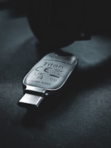 A USB security key marked with the name 'Titan', featuring various certification symbols and model number K40T, lies on a dark surface. The lighting creates a moody atmosphere with emphasis on the metallic texture of the key.