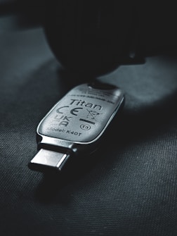 A USB security key marked with the name 'Titan', featuring various certification symbols and model number K40T, lies on a dark surface. The lighting creates a moody atmosphere with emphasis on the metallic texture of the key.