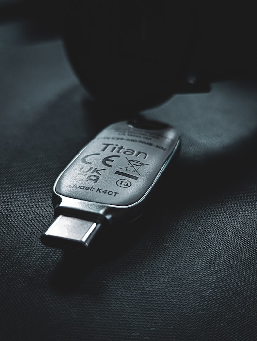 A USB security key marked with the name 'Titan', featuring various certification symbols and model number K40T, lies on a dark surface. The lighting creates a moody atmosphere with emphasis on the metallic texture of the key.
