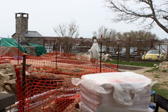A construction site features a partially built stone structure with a chimney, surrounded by orange safety netting. A statue of a robed figure stands in the background, near a row of parked cars. The area is lined with trees and scattered rocks and construction materials suggest ongoing work.