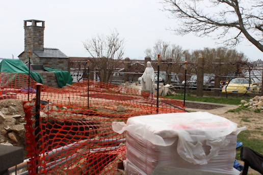 A construction site features a partially built stone structure with a chimney, surrounded by orange safety netting. A statue of a robed figure stands in the background, near a row of parked cars. The area is lined with trees and scattered rocks and construction materials suggest ongoing work.