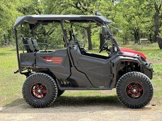 A rugged off-road vehicle with large, knobby tires and a robust black frame stands on a grassy area surrounded by trees. The vehicle features a partial roof, open sides, and a striking red accent on the front.