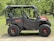 A rugged off-road vehicle with large, knobby tires and a robust black frame stands on a grassy area surrounded by trees. The vehicle features a partial roof, open sides, and a striking red accent on the front.