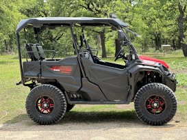 A rugged off-road vehicle with large, knobby tires and a robust black frame stands on a grassy area surrounded by trees. The vehicle features a partial roof, open sides, and a striking red accent on the front.