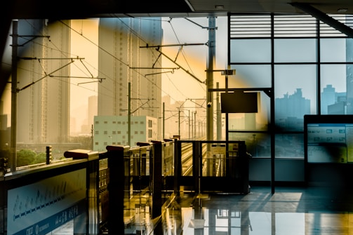 A modern train station platform with sleek glass walls overlooking a cityscape of tall buildings. Sunlight creates a warm, golden glow, casting shadows and reflections on the shiny tiled floor. Overhead cables and tracks are visible, suggesting an urban railway network.