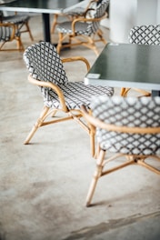 A group of chairs with a woven pattern and bamboo-style frames arranged around black tables. The floor is concrete, and the setting appears to be a casual indoor cafe or outdoor patio.