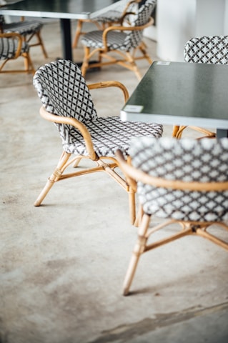 A group of chairs with a woven pattern and bamboo-style frames arranged around black tables. The floor is concrete, and the setting appears to be a casual indoor cafe or outdoor patio.