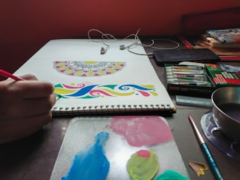 A person is creating colorful mandala art on a sketchbook. The workspace features various art supplies including paint tubes, a palette with mixed colors, brushes, and a cup possibly used for water. Earphones are also laid out on the desk.