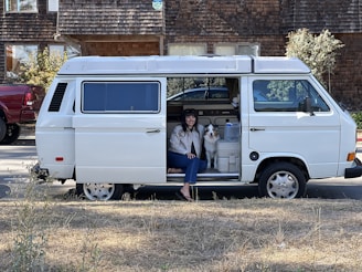 A woman and a dog are sitting inside a white van parked on the side of the road. The van door is open, revealing the interior and its contents, including a water bottle and some storage containers. In the background, there is a wooden house with a shingled exterior.