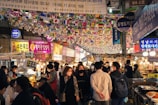 A bustling indoor marketplace with numerous people strolling through narrow pathways lined with food stalls. The ceiling is adorned with multiple small national flags hanging from strings. Bright neon signs and banners with Korean text are visible, enhancing the lively and vibrant atmosphere.