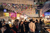 A bustling indoor marketplace with numerous people strolling through narrow pathways lined with food stalls. The ceiling is adorned with multiple small national flags hanging from strings. Bright neon signs and banners with Korean text are visible, enhancing the lively and vibrant atmosphere.