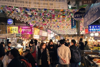 A bustling indoor marketplace with numerous people strolling through narrow pathways lined with food stalls. The ceiling is adorned with multiple small national flags hanging from strings. Bright neon signs and banners with Korean text are visible, enhancing the lively and vibrant atmosphere.