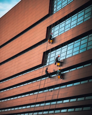 Several workers are suspended on ropes, cleaning or maintaining the windows of a large, modern building with brown panels and blue-tinted glass. The structure has a slightly curved facade, and the workers are equipped with protective helmets and gear.