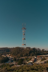 A tall radio tower stands prominently against a clear blue sky, situated on a hill covered with dense green trees. At the base of the hill, a suburban area with numerous houses is visible, suggesting an urban setting at the edge of a forested region.