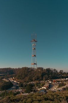A tall radio tower stands prominently against a clear blue sky, situated on a hill covered with dense green trees. At the base of the hill, a suburban area with numerous houses is visible, suggesting an urban setting at the edge of a forested region.