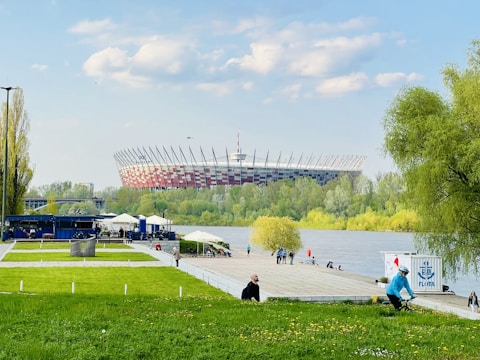 A large soccer stadium with a unique architectural design is situated in the background, surrounded by lush greenery and a river in the foreground. People are enjoying the outdoor space; some are walking along the path, while others are cycling. The sky is clear with a few clouds.
