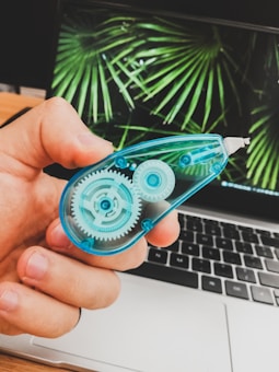 A hand is holding a correction tape dispenser with visible gears inside a transparent blue casing. In the background, a laptop is open displaying a wallpaper with green palm leaves.