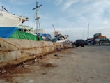 A dock area with moored ships and cranes in the background. The dock is lined with large sandbags, and there are various industrial elements like a truck and containers visible. The setting appears busy, with several shipping-related activities likely taking place.
