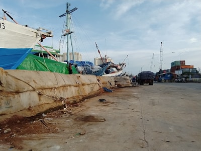 A dock area with moored ships and cranes in the background. The dock is lined with large sandbags, and there are various industrial elements like a truck and containers visible. The setting appears busy, with several shipping-related activities likely taking place.