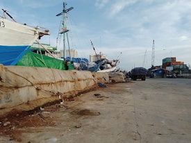 A dock area with moored ships and cranes in the background. The dock is lined with large sandbags, and there are various industrial elements like a truck and containers visible. The setting appears busy, with several shipping-related activities likely taking place.