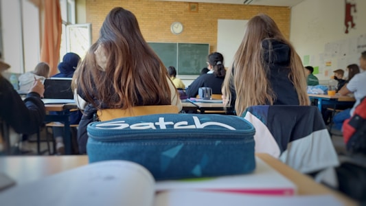 A classroom setting with students sitting at desks facing a blackboard. A pencil case labeled 'Satch' is prominently placed on the desk in the foreground. The classroom is filled with natural light from large windows on the side, and a clock is visible on the wall above the blackboard.