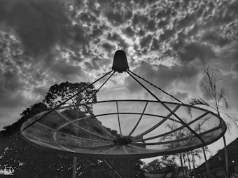 A large satellite dish silhouette against a dramatic sky filled with textured clouds. Trees and palm plants are visible in the background, framing the dish and adding depth to the composition.