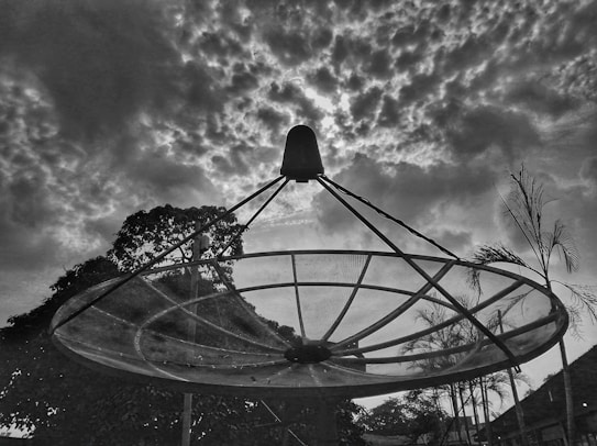 A large satellite dish silhouette against a dramatic sky filled with textured clouds. Trees and palm plants are visible in the background, framing the dish and adding depth to the composition.