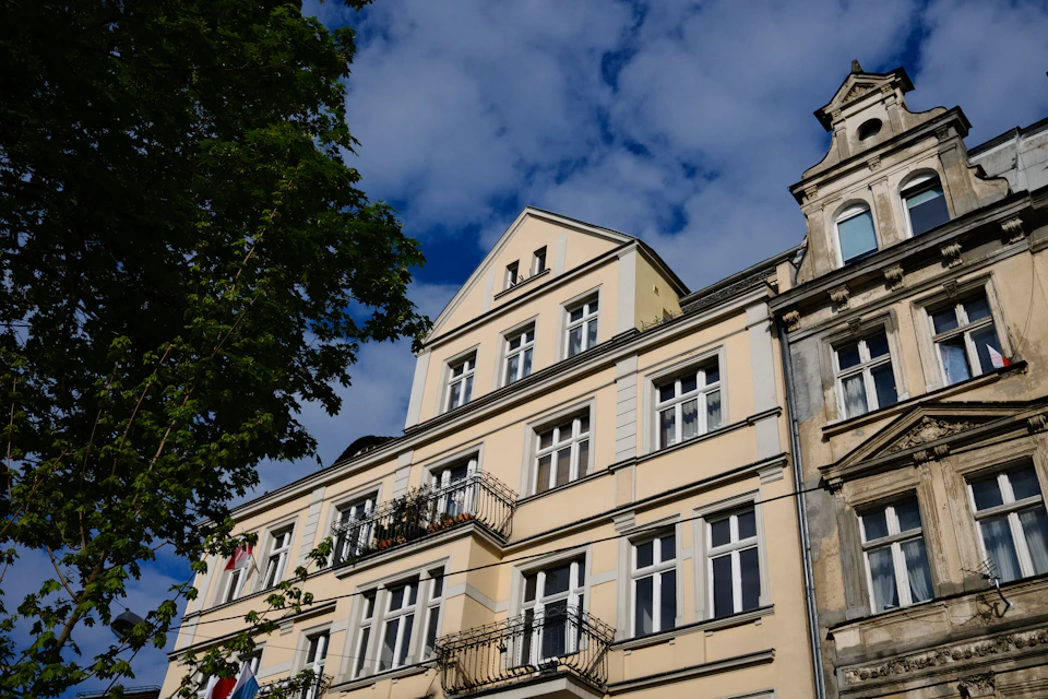 The image depicts a two-story building with a yellow facade and white trim, situated on a street corner. The building features a balcony on the second floor, adding an element of architectural interest to its design. A tree can be seen in front of the building, providing some natural greenery to the urban setting.
