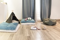 A cozy room setup featuring a small black tent with a soft cushion inside, several plush blue pillows on the floor, a pet bed, a white food bowl with a chew toy inside, and a hedgehog-shaped toy sitting on wooden flooring. The space is brightened by natural light coming through sheer curtains.