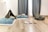 A cozy room setup featuring a small black tent with a soft cushion inside, several plush blue pillows on the floor, a pet bed, a white food bowl with a chew toy inside, and a hedgehog-shaped toy sitting on wooden flooring. The space is brightened by natural light coming through sheer curtains.