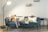 Two small dogs wearing blue bandanas are sitting on a teal velvet couch with several pillows. A floor lamp is positioned to the left, and a modern black side table is on the right. The background features a light-colored wall with small decorative shelves.