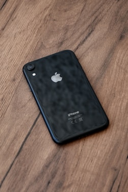A black smartphone with an Apple logo is placed on a wooden surface, displaying its back panel. The phone's camera and flash are visible at the upper corner.