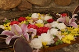 A variety of colorful flowers, including pink lilies, red roses, white carnations, and yellow daisies, are artistically arranged and floating in water. The background features a rustic stone wall, adding a natural element to the floral arrangement.