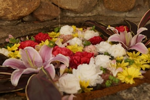 A variety of colorful flowers, including pink lilies, red roses, white carnations, and yellow daisies, are artistically arranged and floating in water. The background features a rustic stone wall, adding a natural element to the floral arrangement.